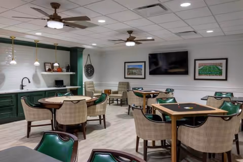A well-lit common area with multiple tables and chairs arranged for socializing or games. The room features green cabinetry with a white countertop and backsplash, three pendant lights, ceiling fans with lights, a wall clock, framed pictures, and a large flat-screen TV mounted on the wall.