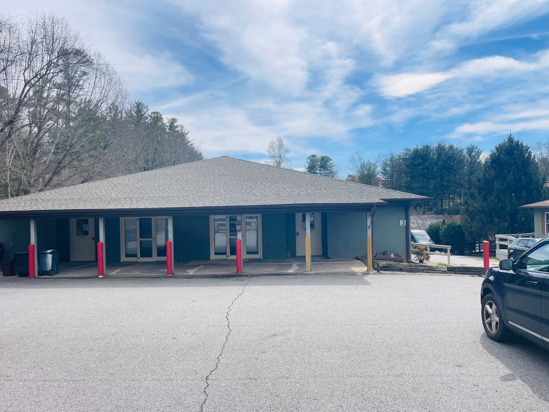 Exterior view of a single-story building with a sloped roof, several doors and windows, red protective bollards in front, a parked black car on the right side, and trees in the background under a partly cloudy sky.