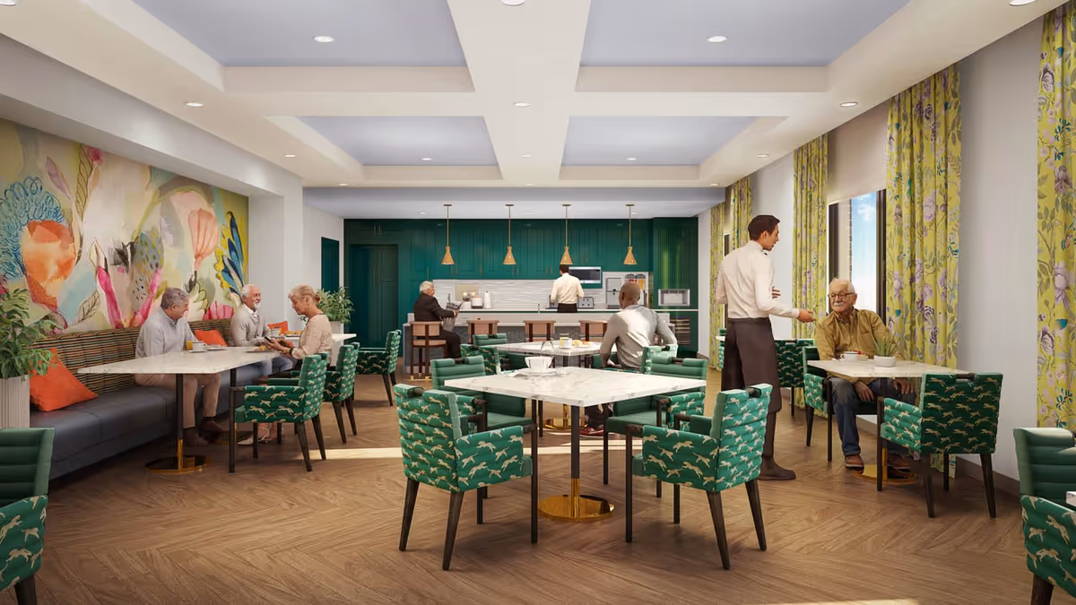 A bright and modern dining area in a senior living facility with several elderly people seated at tables and a waiter serving one of them. The room features green patterned chairs, marble-top tables, a colorful mural on one wall, large windows with floral curtains, and a kitchen area with green cabinetry and pendant lights in the background.