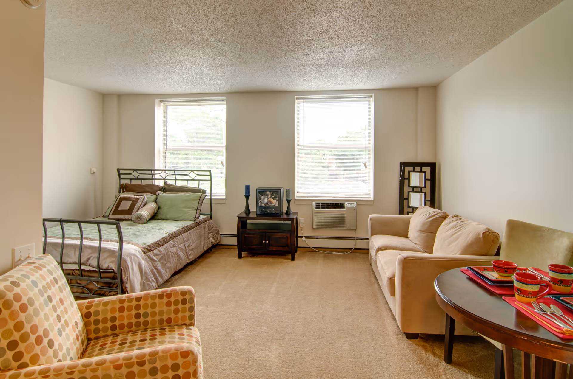 A senior living facility room with a bed on the left side, a small dark wooden table with decorative items between two windows, a beige couch on the right, and a round table with colorful cups and placemats in the foreground. There is also a patterned armchair in the front left corner.