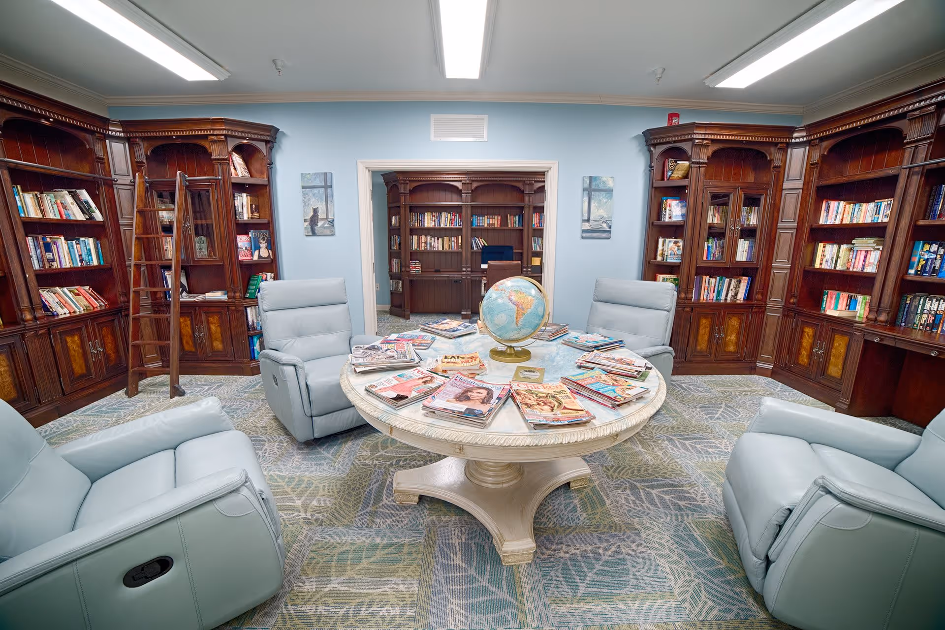 A cozy library room with light blue walls and carpet featuring a leaf pattern. The room has dark wooden bookshelves filled with books along the walls and a rolling ladder on the left side. In the center, there is a round white table with a globe and several magazines spread out on top. Surrounding the table are four light blue recliner chairs. Through an open doorway, another room with more bookshelves and a desk is visible.
