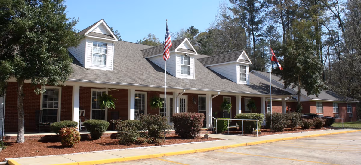 Exterior view of a brick and white siding senior living facility with flags and landscaping.