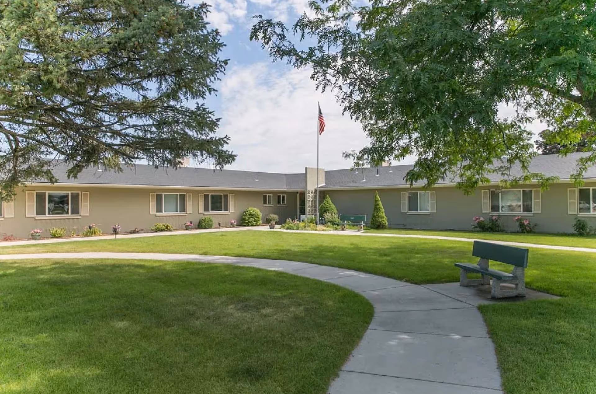 Outdoor view of River's Edge Rehabilitation & Living Center showing a single-story building with multiple windows, a well-maintained lawn, a curved concrete walkway, two benches, large trees providing shade, and an American flag on a flagpole in the center.