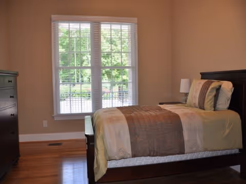Sunlit bedroom with a single bed, nightstand and lamp beside a large window overlooking trees.