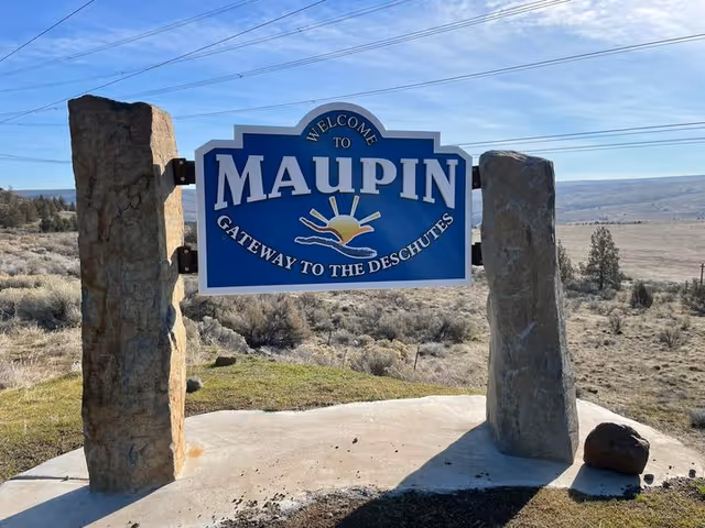 A blue and white welcome sign for Maupin, labeled as the Gateway to the Deschutes, mounted between two large stone pillars with a natural landscape and clear sky in the background.