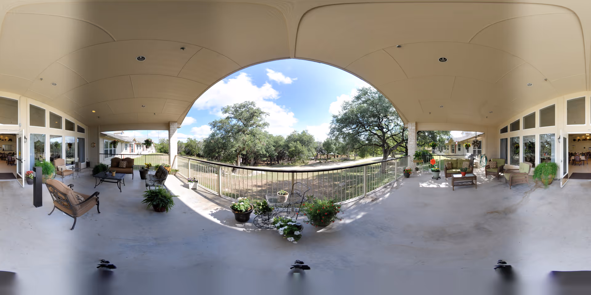 Covered outdoor patio area at Heritage Place of Boerne with seating arrangements including chairs and tables, potted plants along the railing, and a view of trees and open land under a partly cloudy sky.
