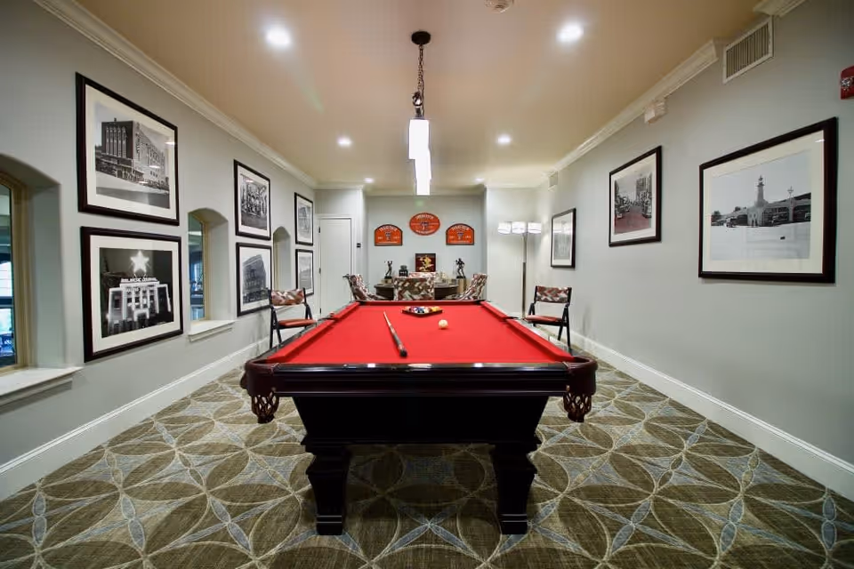 Interior room with a red felt pool table centered in the space, surrounded by chairs and decorated with framed black and white photographs on the walls. The room has patterned carpet flooring and a modern overhead light fixture above the pool table.