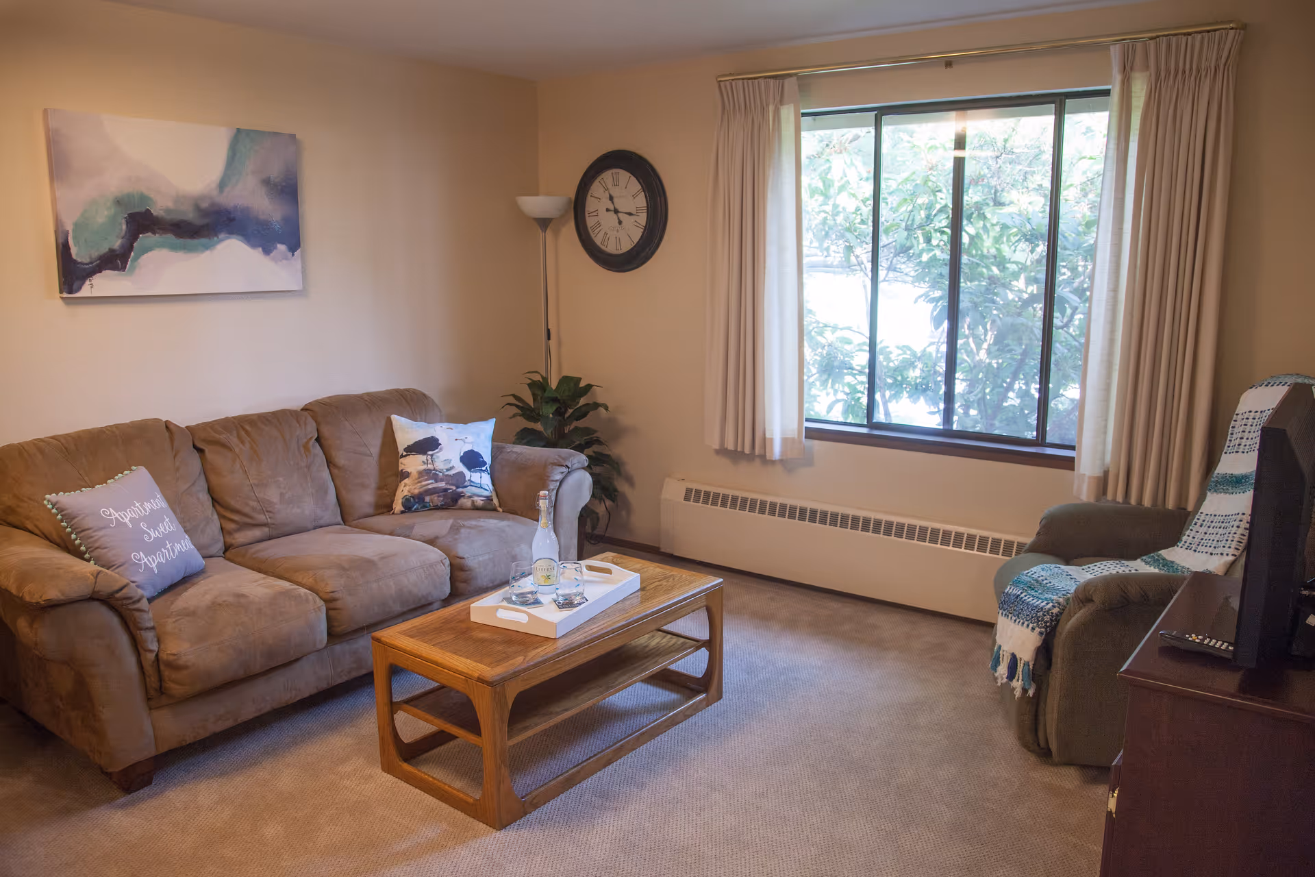 A cozy living room with a brown couch adorned with decorative pillows, a wooden coffee table with a white tray holding a bottle and two glasses, a green armchair with a blue and white throw blanket, a large window with cream curtains, a wall clock, a floor lamp, a potted plant, and a piece of abstract wall art.