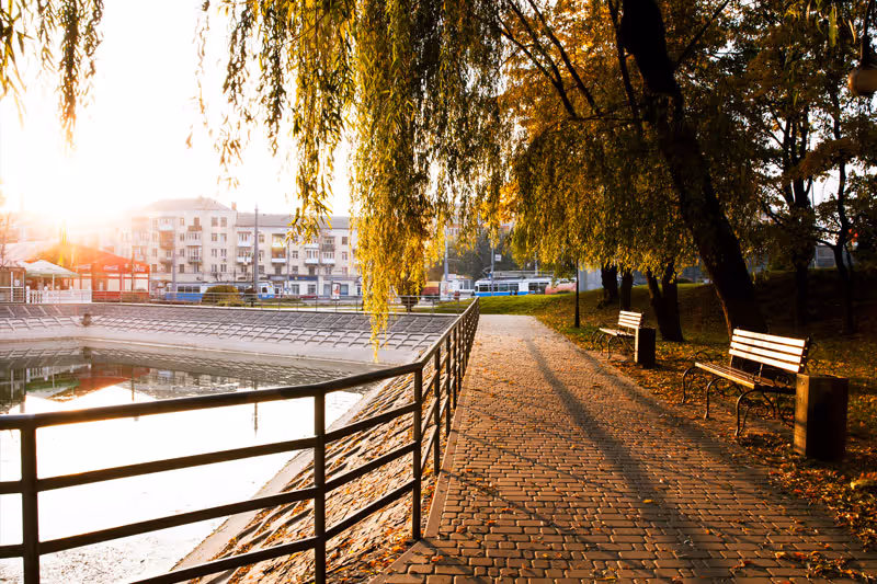 A serene outdoor pathway along a body of water with a metal railing on the left and benches on the right under large trees with hanging branches. The sun is setting, casting long shadows and a warm golden light over the scene. Buildings and a street with vehicles are visible in the background.