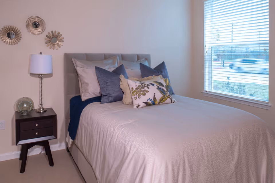 Neatly made bed with decorative pillows, a nightstand with a lamp and decor, and a window with blinds.