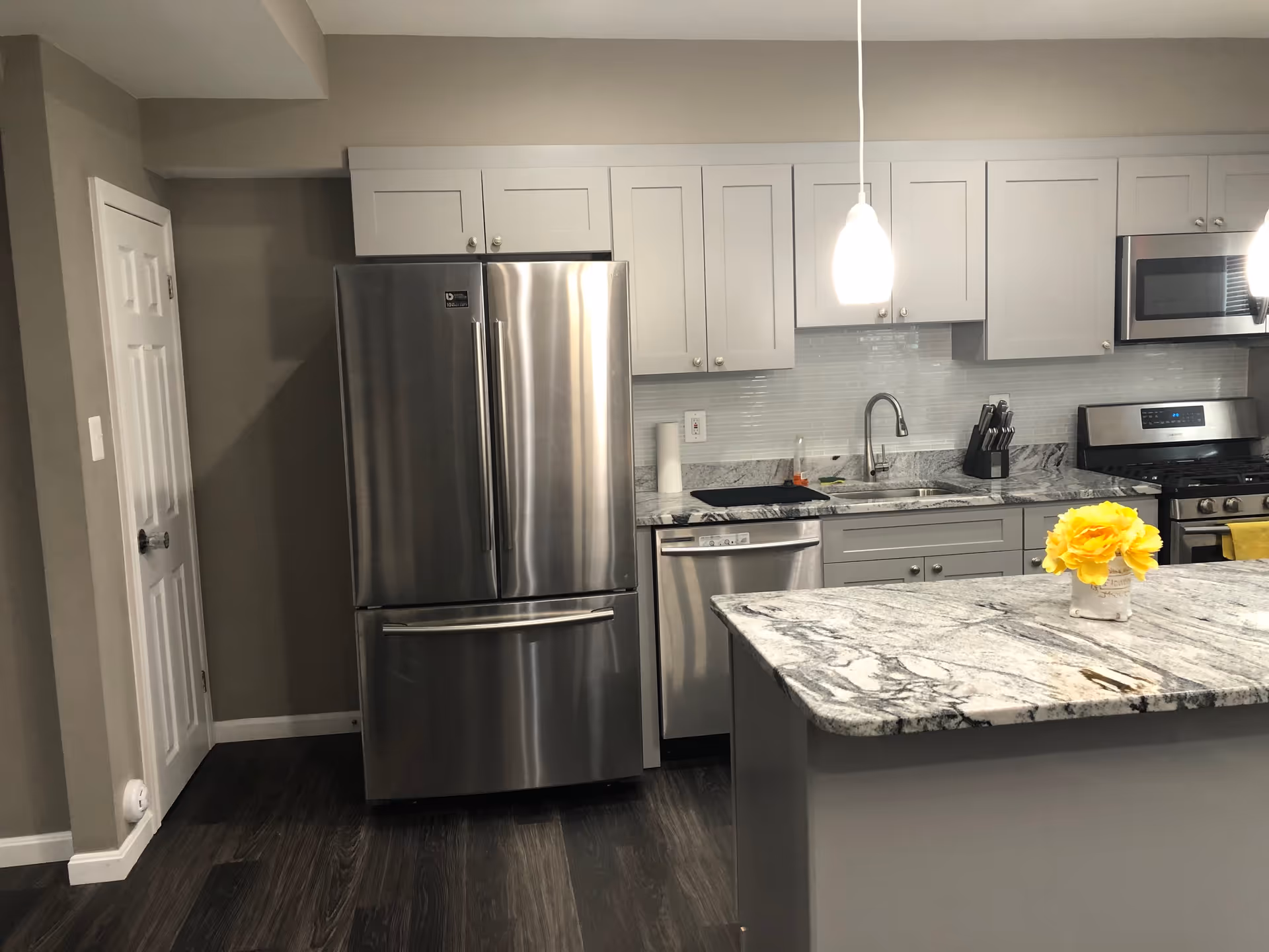 Modern kitchen with stainless steel refrigerator, dishwasher, microwave, and stove. White cabinets and a marble countertop island with a small vase of yellow flowers. Two pendant lights hang above the island.