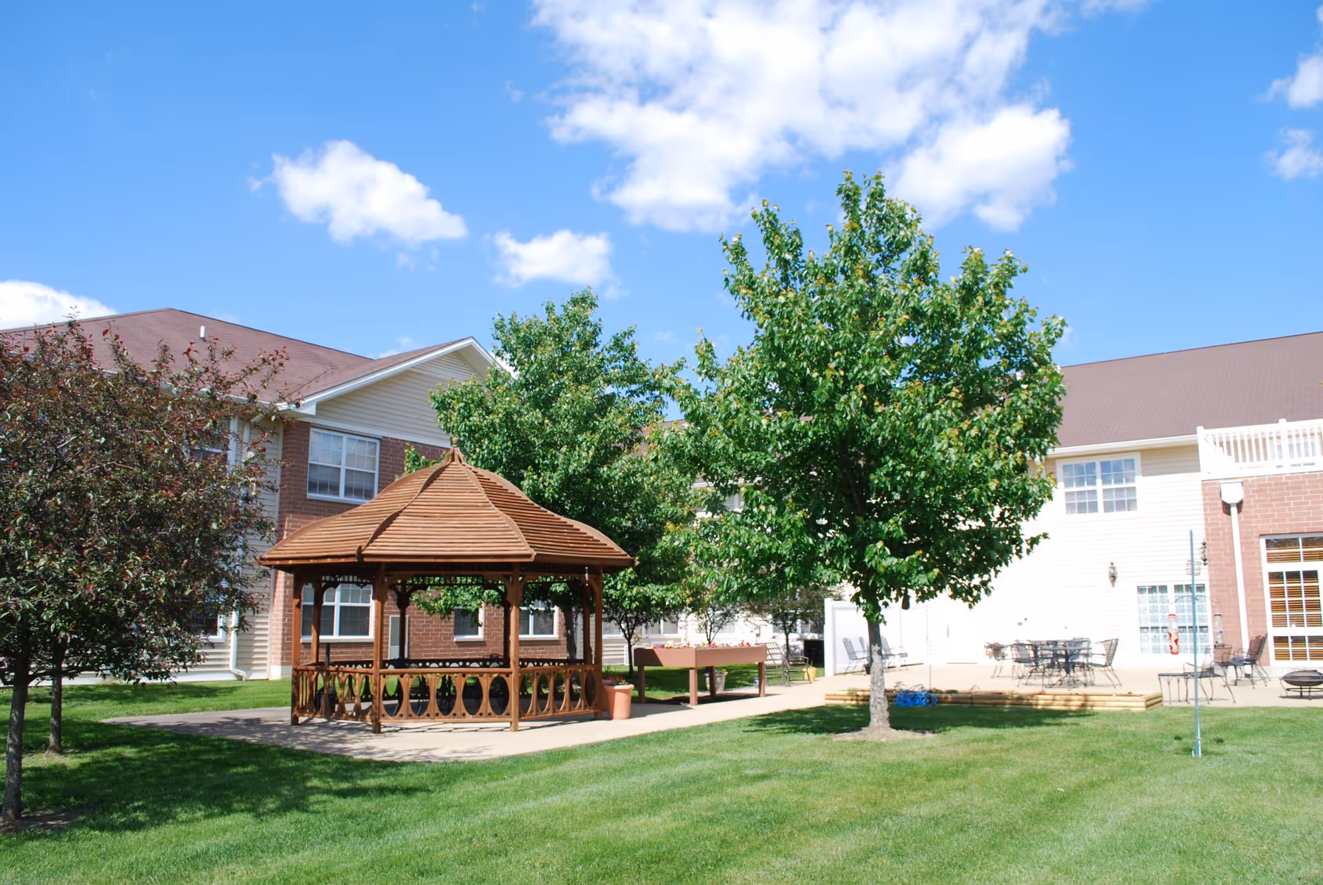 Outdoor area of Bowman Estates of Danville featuring a wooden gazebo, green grass, several trees, and a patio with tables and chairs under a blue sky with some clouds.