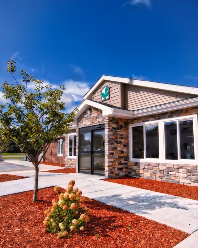 Front entrance of a single-story assisted living building with a stone facade, paved walkway, small tree and landscaped flower beds under a blue sky.