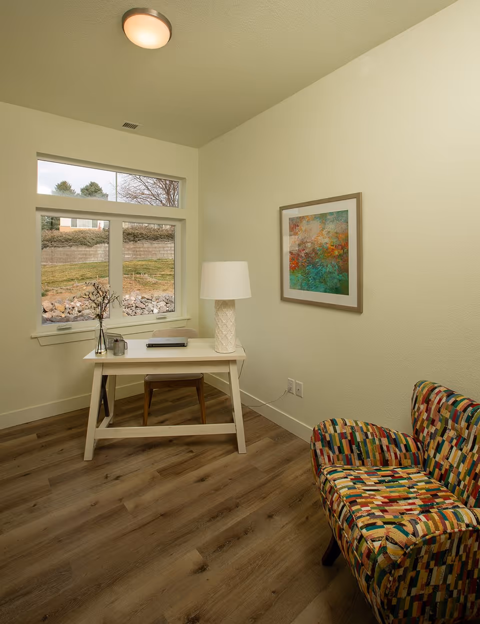 A small room with a wooden desk and chair near a window showing an outdoor view. On the desk are a lamp, a laptop, and a small vase with decorative branches. A colorful patterned armchair is positioned to the right, and a framed abstract painting hangs on the wall.