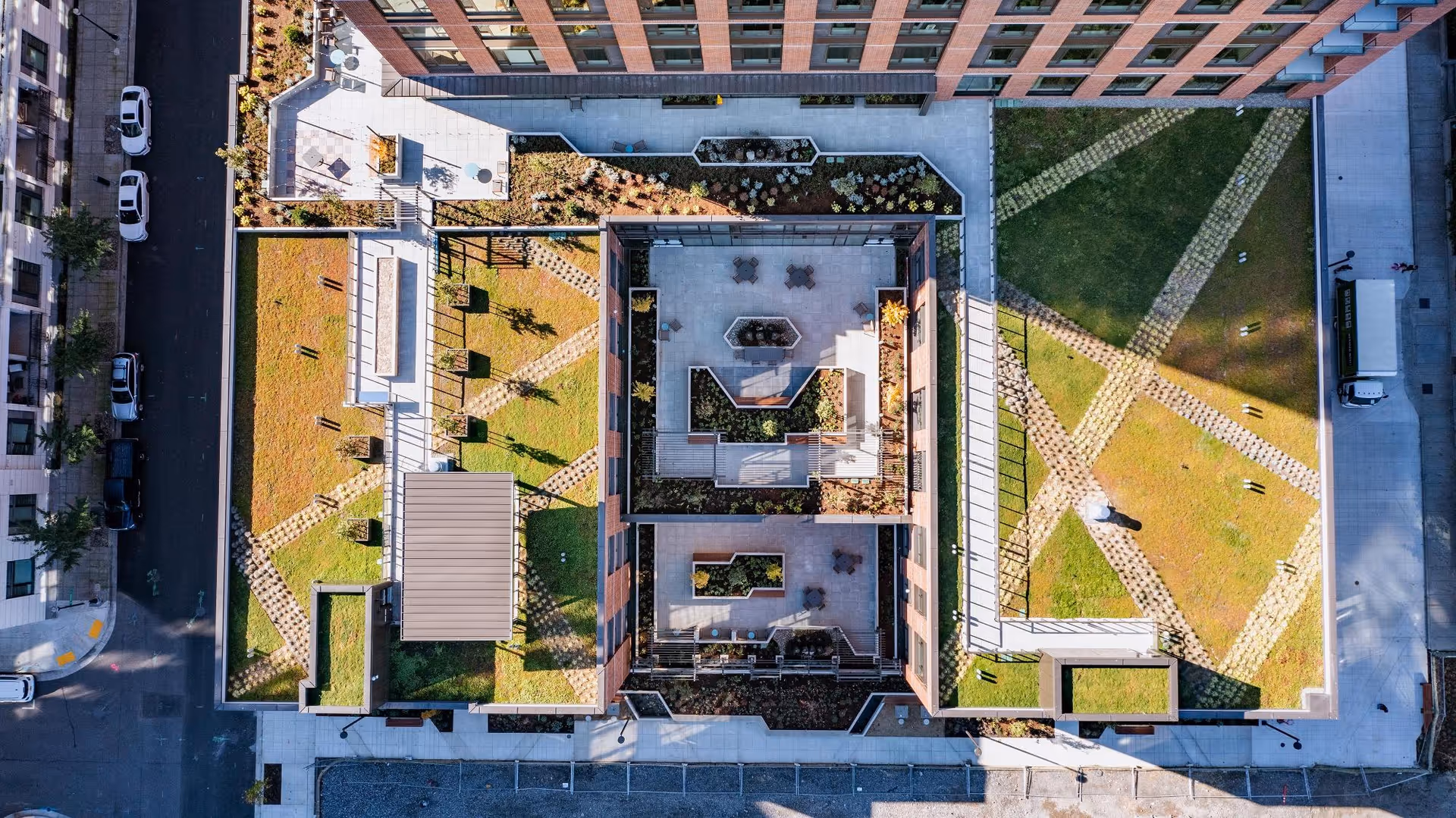Aerial view of a building rooftop garden with green grass, paved walkways, seating areas, and planters. The rooftop is divided into multiple sections with geometric patterns and surrounded by the building structure and adjacent streets with parked cars.
