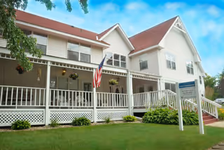 Exterior view of a two-story white building with a red roof, a covered porch with white railings, hanging flower pots, an American flag, and a sign on the lawn. The sky is blue with some clouds.