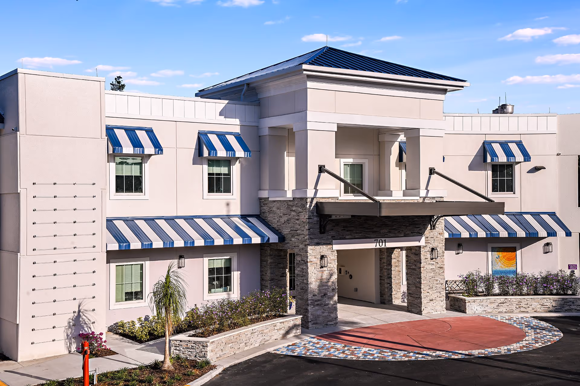 Exterior view of a two-story building with beige walls and blue and white striped awnings over the windows and entrance. The entrance features stone pillars and a covered driveway with the number 701 above it. There are landscaped areas with plants and flowers around the building under a clear blue sky.