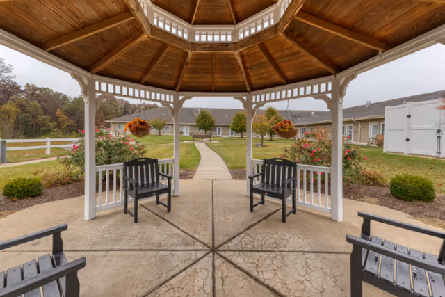View from inside a wooden gazebo with a peaked roof, looking out onto a paved walkway leading to a single-story building surrounded by green lawns, bushes, and trees. Four black chairs are placed inside the gazebo, and hanging flower baskets are visible on the gazebo's posts.