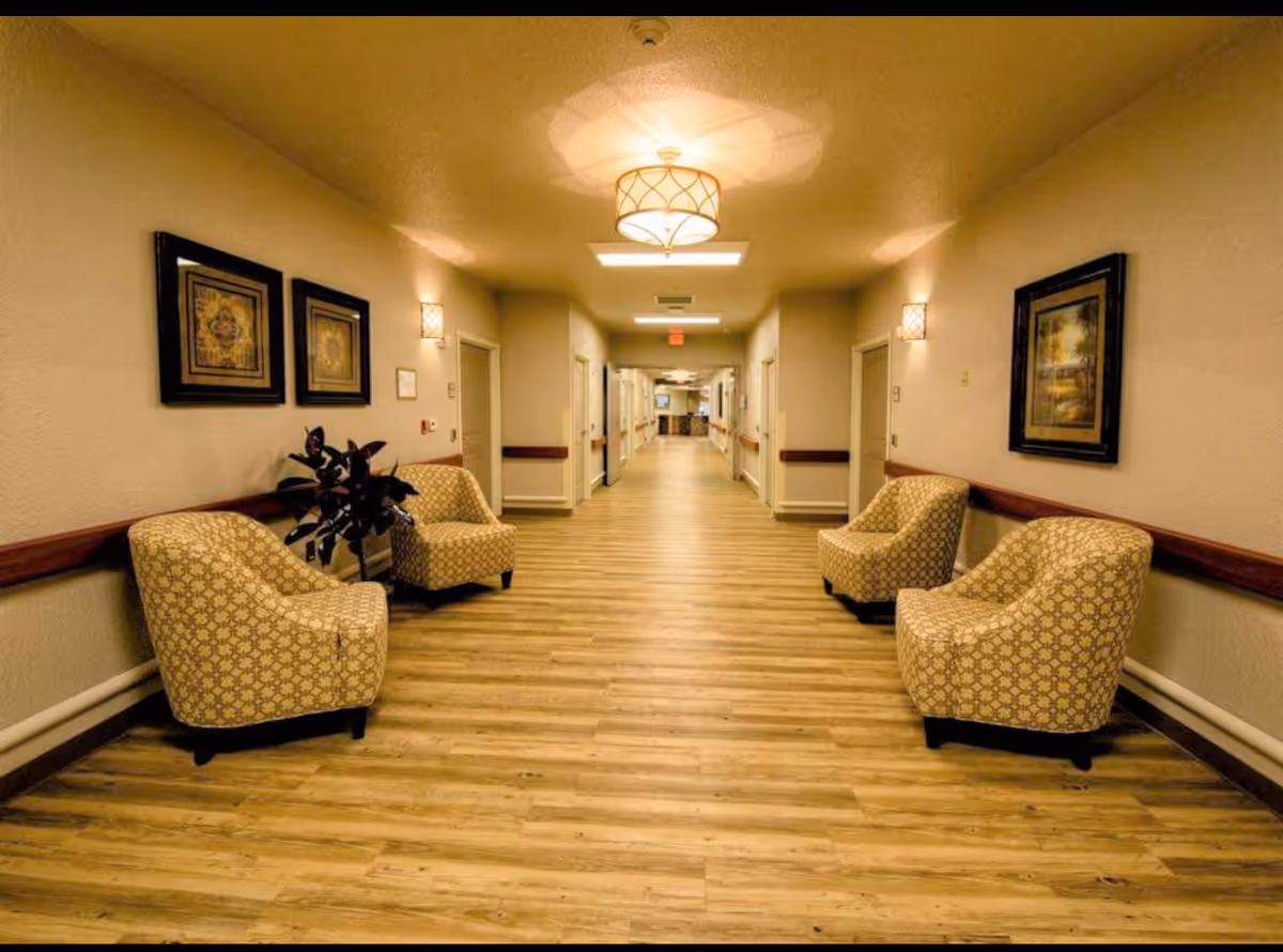 A well-lit hallway in a senior care facility with wooden flooring, beige walls, and four patterned armchairs arranged in pairs along the walls. The hallway is decorated with framed artwork and has ceiling lights and wall sconces providing warm lighting.