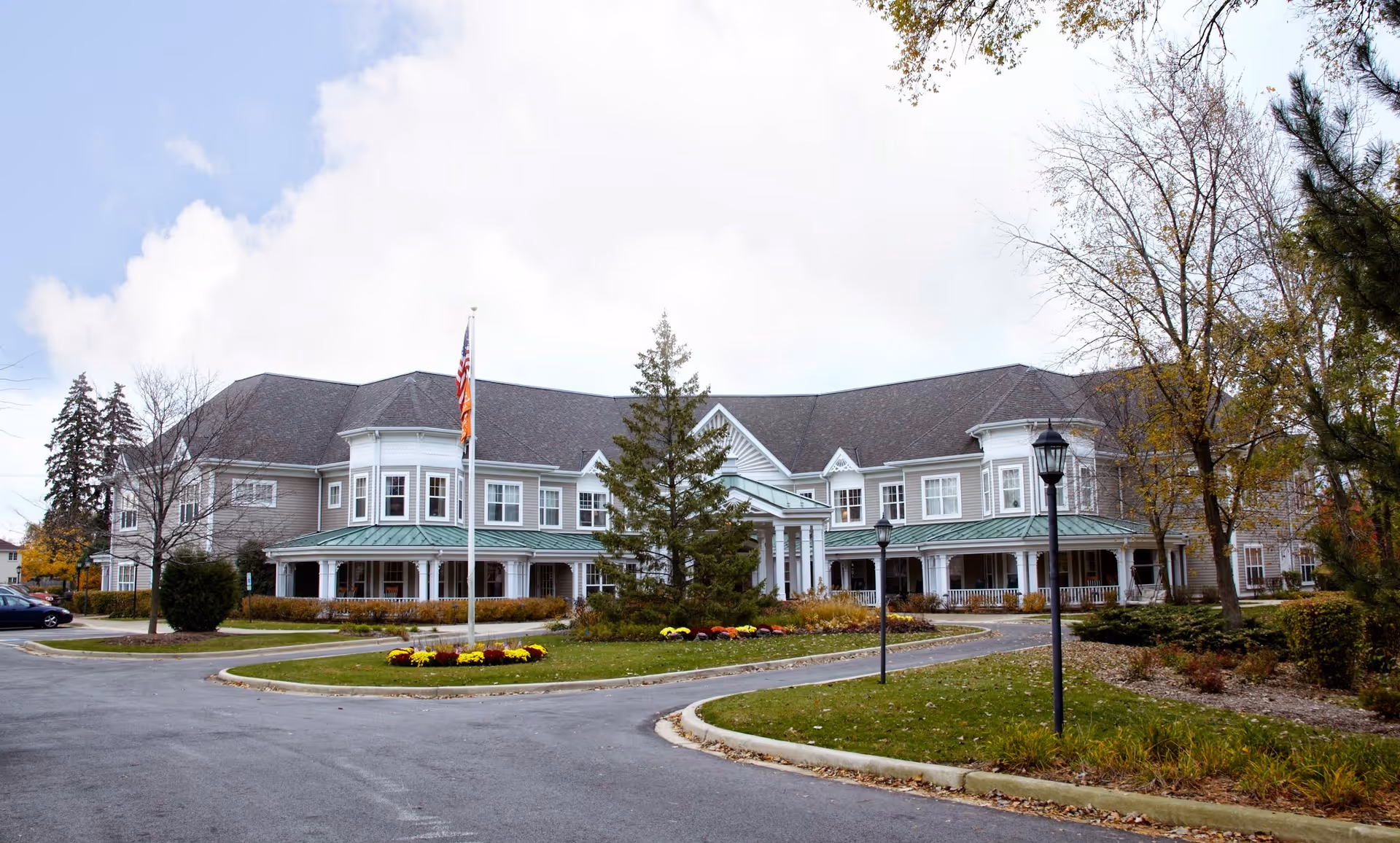 Exterior view of a large two-story senior living facility building with a circular driveway, landscaped greenery, and an American flag on a flagpole in front. The building has multiple windows, a green roof over the porch area, and trees surrounding the property.