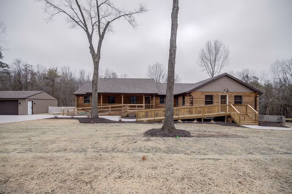Front view of a single-story log-style building with a long wheelchair ramp, detached garage, and bare trees on a cloudy day.