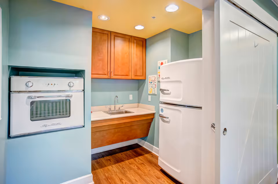 Small kitchen area with a vintage white oven built into a teal wall, wooden cabinets above a countertop with a sink, and a retro white refrigerator. The floor is wooden, and the ceiling is painted yellow with recessed lighting.