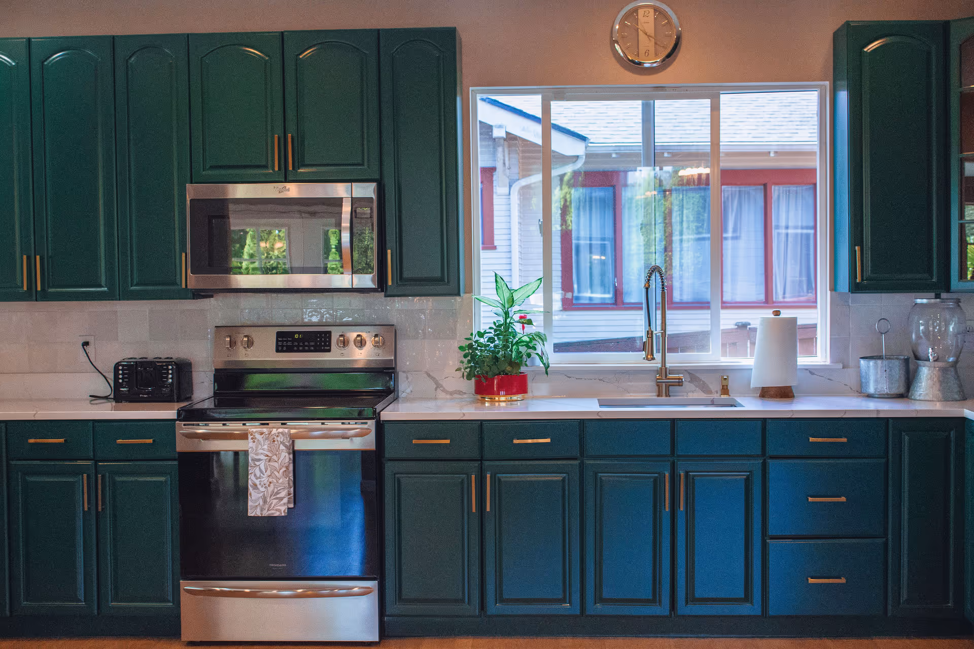 A bright kitchen with dark green cabinets, stainless steel stove and microwave, a sink under a large window, and countertop accessories.