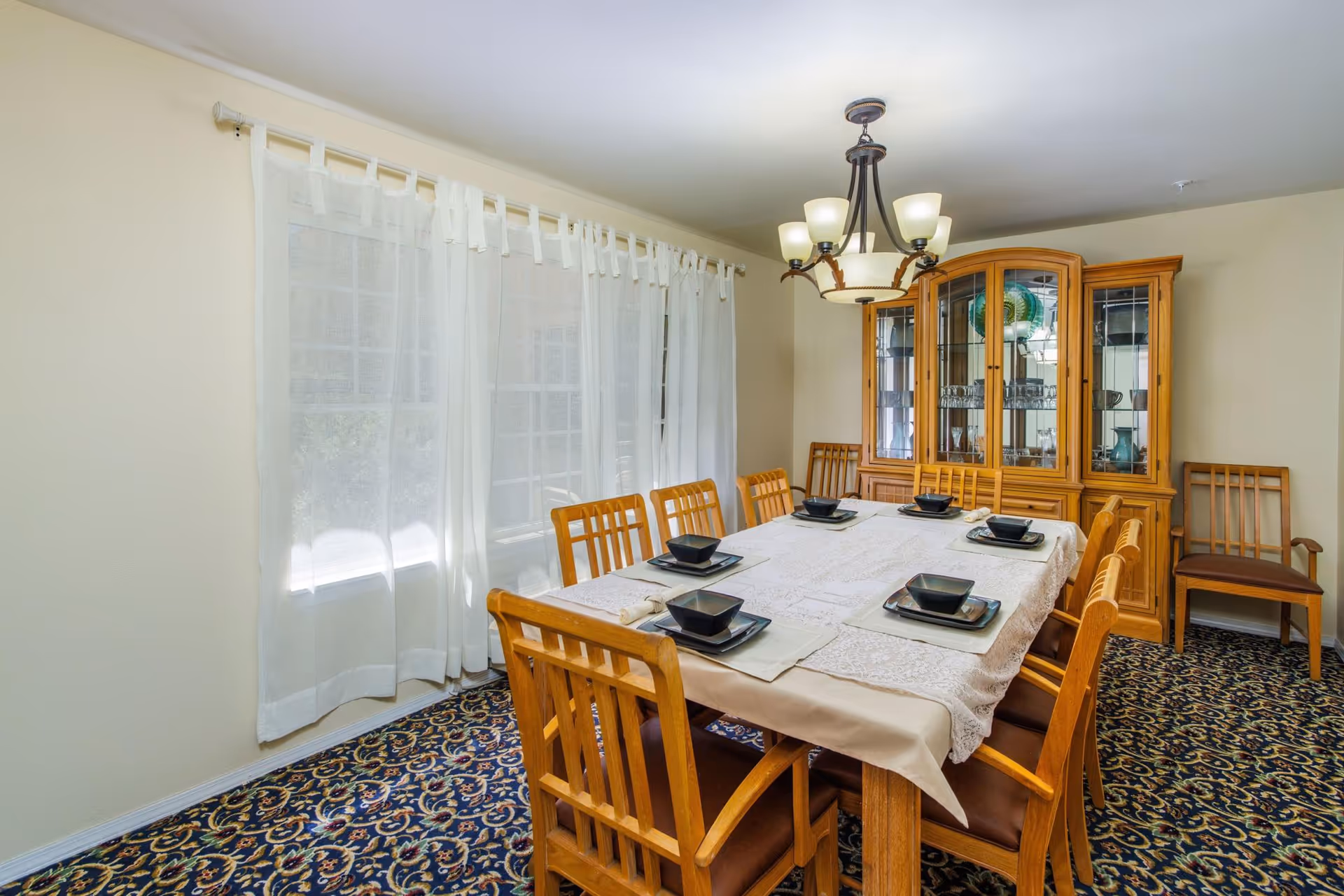 Dining room with a long wooden table set for eight, wooden chairs, a china cabinet, chandelier, and sheer curtains covering windows.