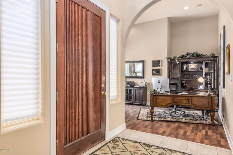 View of an office area from an entryway with a large wooden door on the left. The office features a wooden desk with a computer, a leather office chair, a wooden hutch with decorative items, and a patterned rug on the wooden floor. The walls are light-colored, and there is an arched doorway leading into the office space.