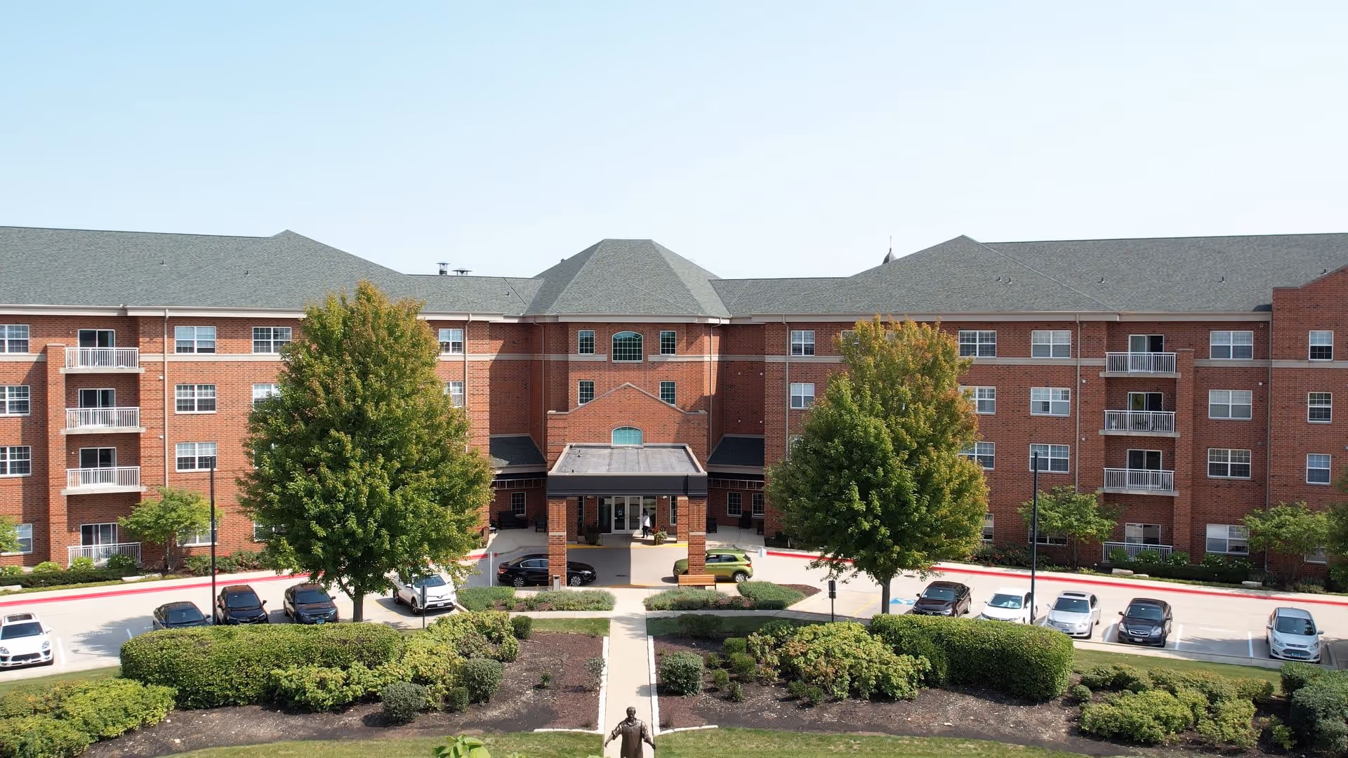 Front exterior view of Villa St. Benedict, a large multi-story brick building with multiple windows and balconies. There is a covered entrance with a driveway and several parked cars. The landscaping includes green bushes, trees, and a walkway leading to the entrance.
