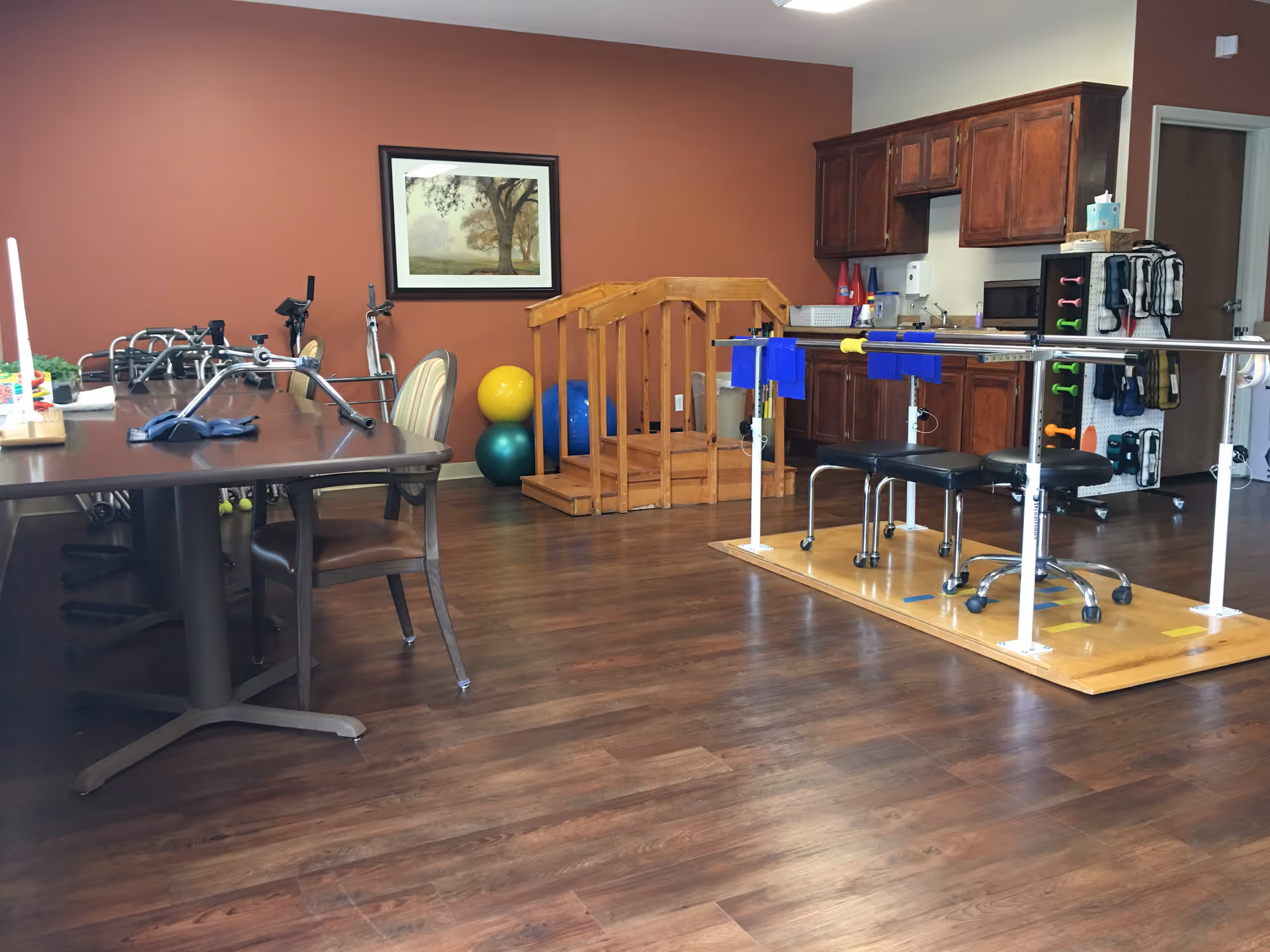A rehabilitation room with wooden flooring, featuring parallel bars for physical therapy, exercise balls, a small wooden ramp, a table with chairs, and a kitchenette with wooden cabinets in the background.