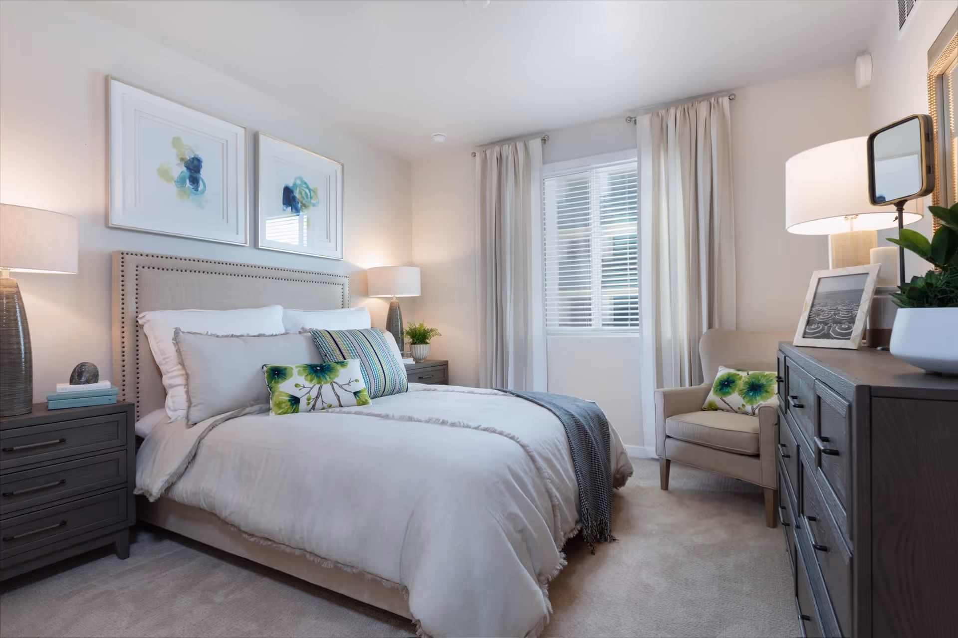 A neatly made bed with beige bedding and decorative pillows in a bright bedroom. The room features two bedside tables with lamps, a window with white blinds and beige curtains, a beige armchair with a floral pillow, and a dark wooden dresser with a lamp, a framed photo, a mirror, and a plant on top. Two abstract paintings hang above the bed.