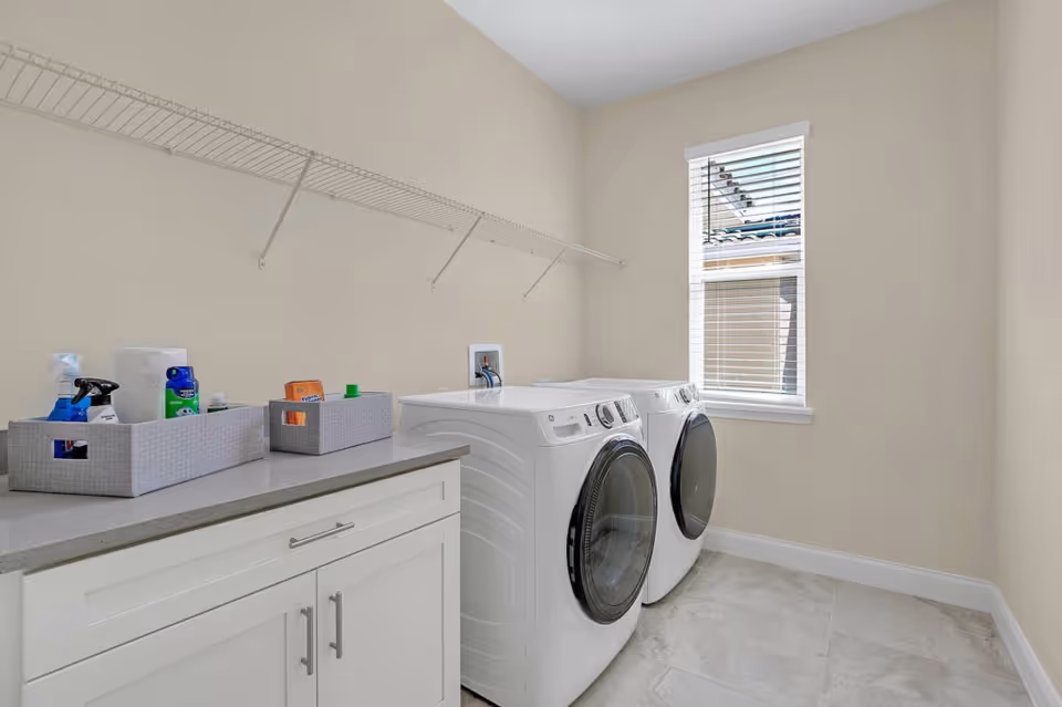A bright laundry room with a front-loading washing machine and dryer side by side, a countertop with two storage baskets containing cleaning supplies, a wire shelf above, and a window with blinds letting in natural light.