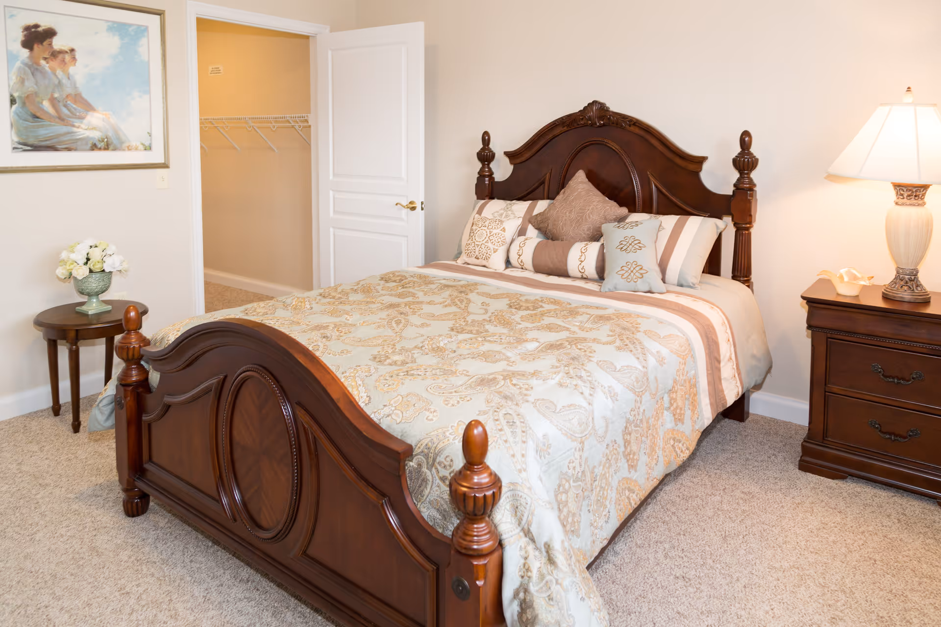 A neatly made bed with a carved wooden headboard and footboard in a bedroom. The bed is covered with a patterned beige and light blue bedspread and several decorative pillows. To the right of the bed is a wooden nightstand with a lamp and a decorative item. To the left is a small round table with a vase of white flowers. An open door reveals a walk-in closet with wire shelving. A framed painting of two women sitting outdoors hangs on the wall.