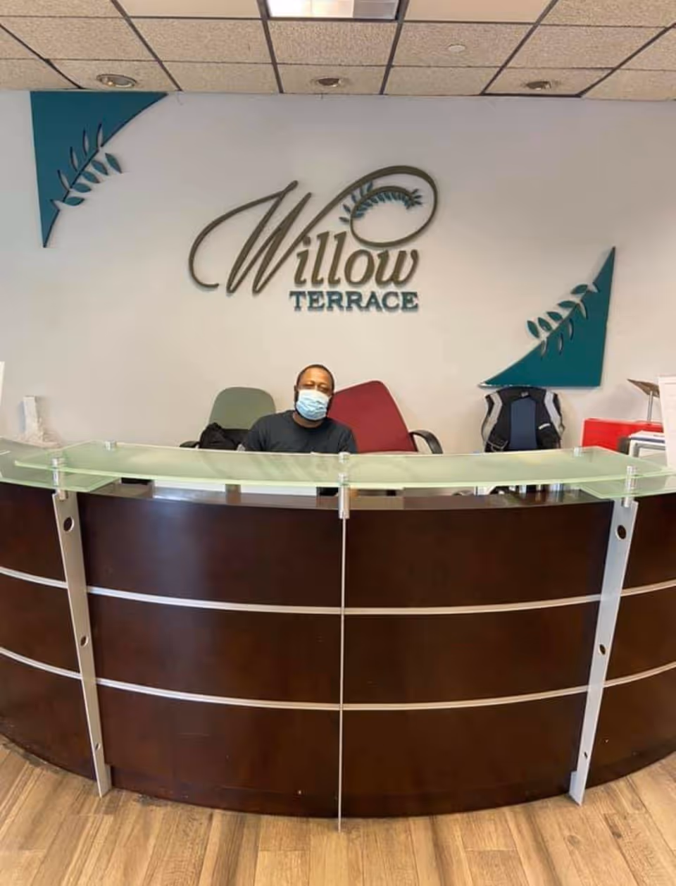 Reception area of Willow Terrace facility with a man wearing a face mask sitting behind a curved dark wood reception desk with a frosted glass top. The wall behind him features the Willow Terrace logo and decorative green corner accents.