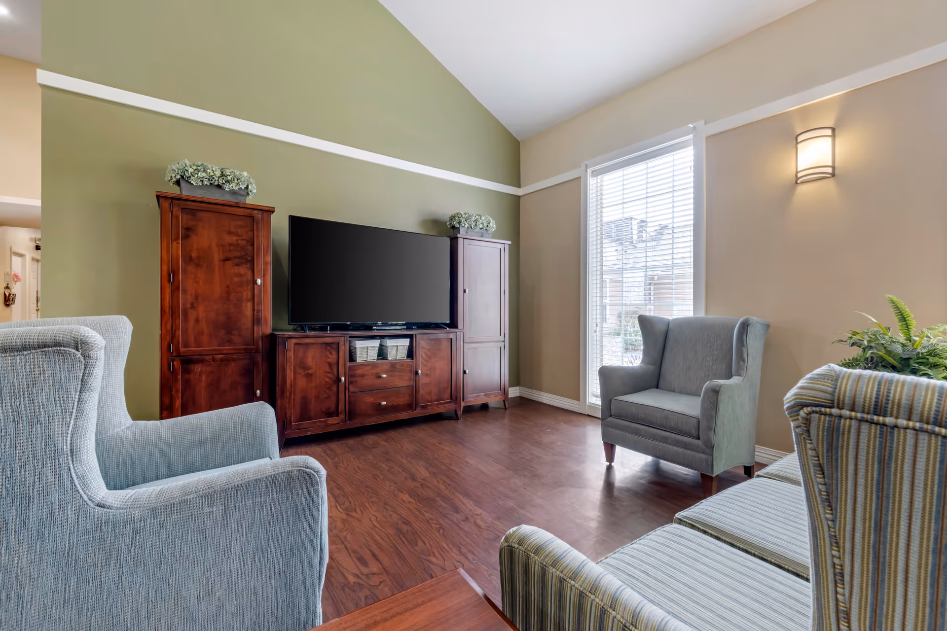 A bright living room with wooden flooring, featuring a large flat-screen TV on a wooden cabinet flanked by two tall wooden cupboards. The room has a green accent wall and beige walls with a window covered by white blinds letting in natural light. There are three upholstered armchairs in different shades of blue and striped fabric arranged around the room, and a wall-mounted light fixture is visible.