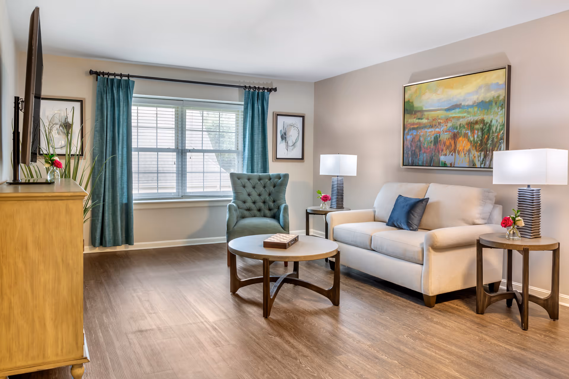 A bright living room with a beige sofa, a blue-green armchair, a round wooden coffee table with a chess/checkers board on it, two side tables with lamps and small flower vases, a large window with teal curtains, and colorful artwork on the walls.