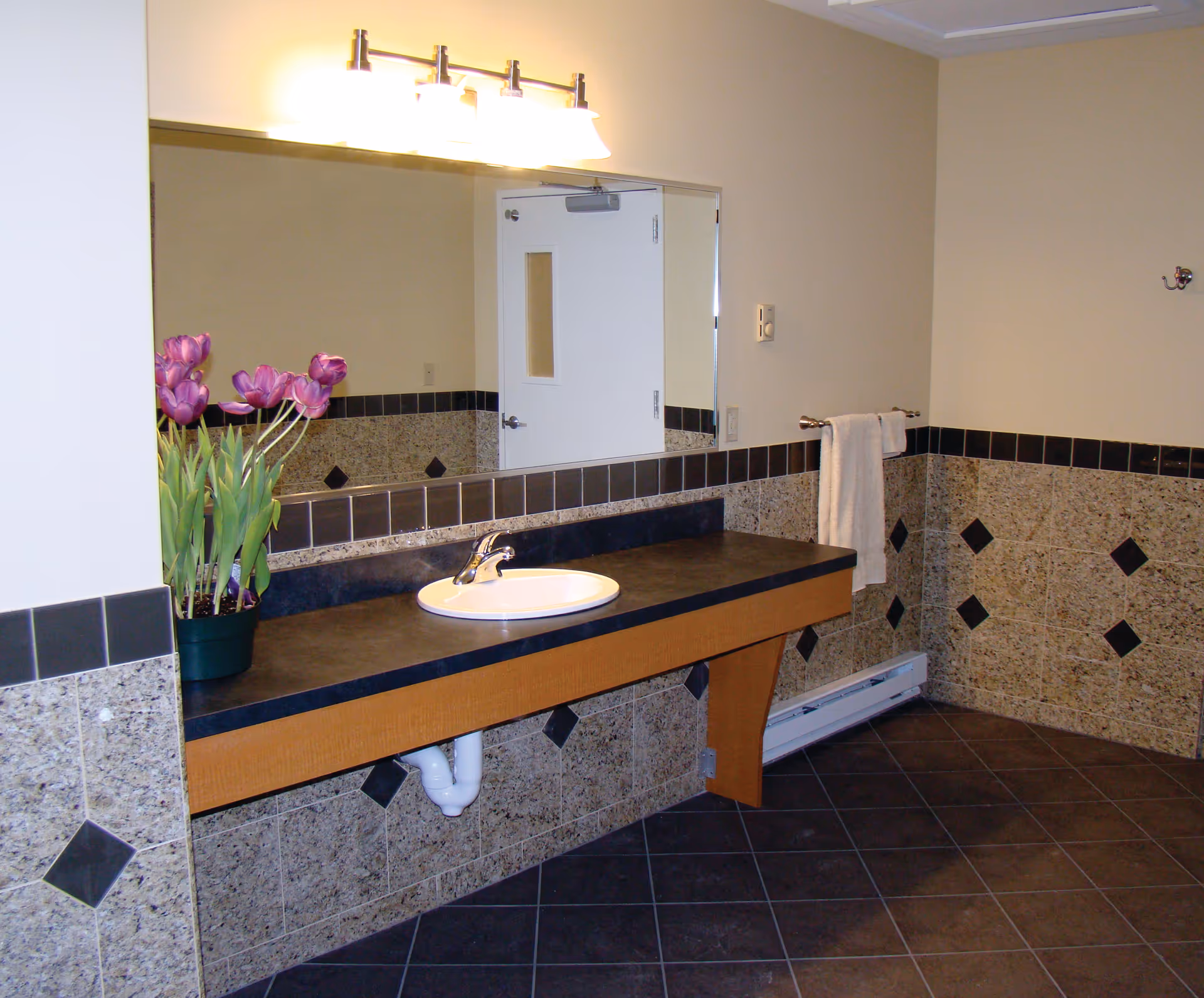 A clean bathroom interior featuring a large mirror above a black countertop with a white sink. There is a potted plant with purple flowers on the left side of the counter. The walls are tiled halfway up with a beige and black diamond pattern, and a white towel hangs on a towel rack on the right wall. The floor is covered with dark tiles, and a white door with a small window is reflected in the mirror.