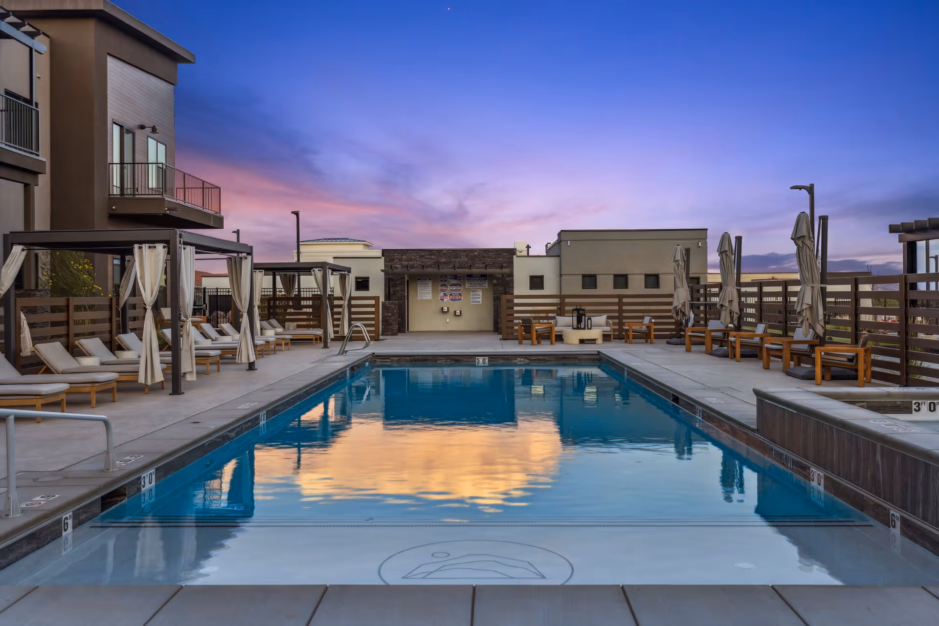 Outdoor swimming pool at sunset with lounge chairs and cabanas on the sides, surrounded by a modern building and fencing under a colorful sky with purple and orange hues.