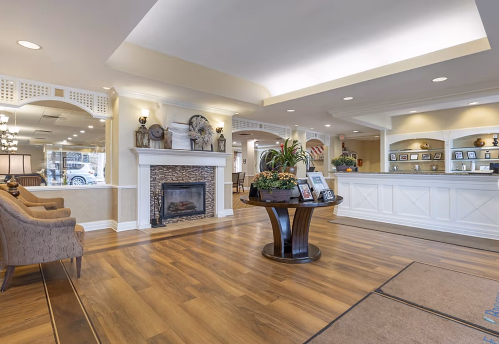 A spacious and well-lit senior living facility lobby with wooden flooring, a round table with plants and framed photos in the center, a white reception desk on the right, a fireplace with decorative items above it on the left, and comfortable chairs along the left wall.