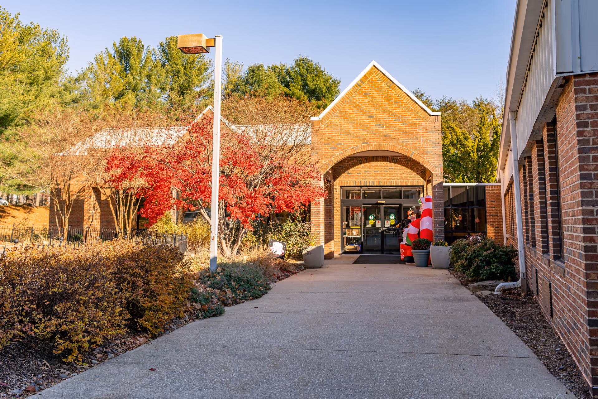 Exterior view of a brick building entrance with a covered walkway, surrounded by trees with autumn foliage and shrubs. There is a concrete pathway leading to the glass double doors, and a festive inflatable decoration near the entrance.