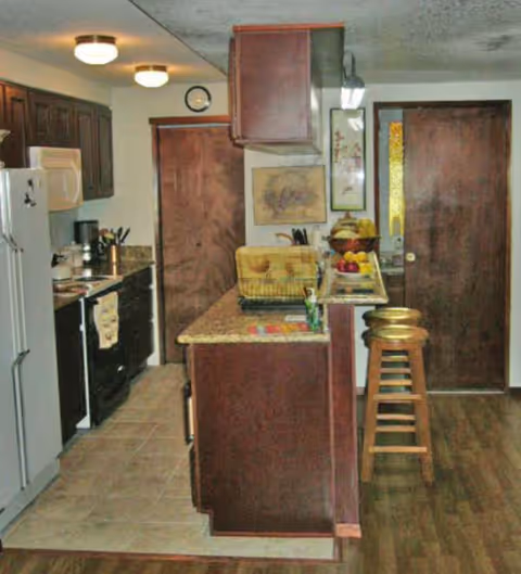 Compact kitchen with island, dark wood cabinets, stove, fridge, and bar stools.