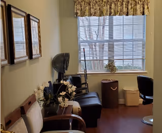 A small waiting or sitting area with chairs along the left wall, a black chair and a hair dryer chair near a window with blinds and a floral valance. There are framed pictures on the wall and a small table with a plant. A trash bin and a white container are near the window.