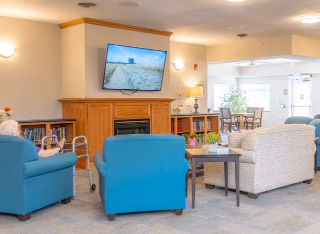 Communal living room with blue and beige armchairs facing a wall-mounted TV above a wooden fireplace, with bookshelves and a side table.