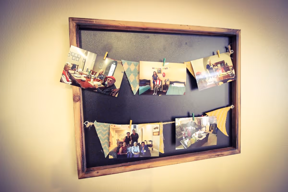 A wooden-framed blackboard hanging on a wall with photos and decorative fabric flags clipped to strings across it. The photos show various indoor scenes with people and furniture, suggesting a warm, communal environment.