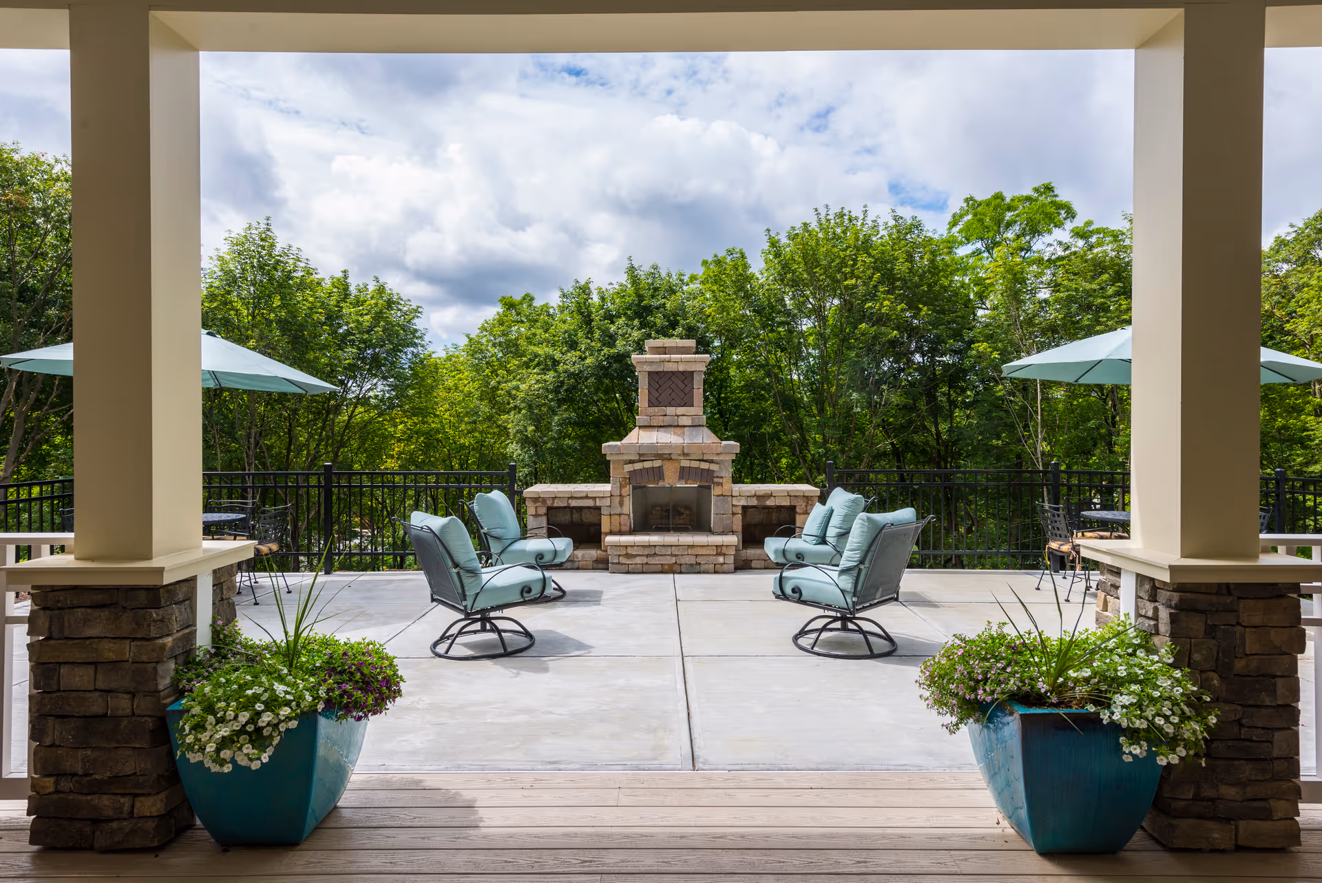 Outdoor patio area with a stone fireplace in the center, surrounded by four cushioned chairs with metal frames. Two large blue planters with flowers are placed on either side of the covered porch entrance. There are also two patio tables with umbrellas on either side of the fireplace. Green trees and a partly cloudy sky are visible in the background.