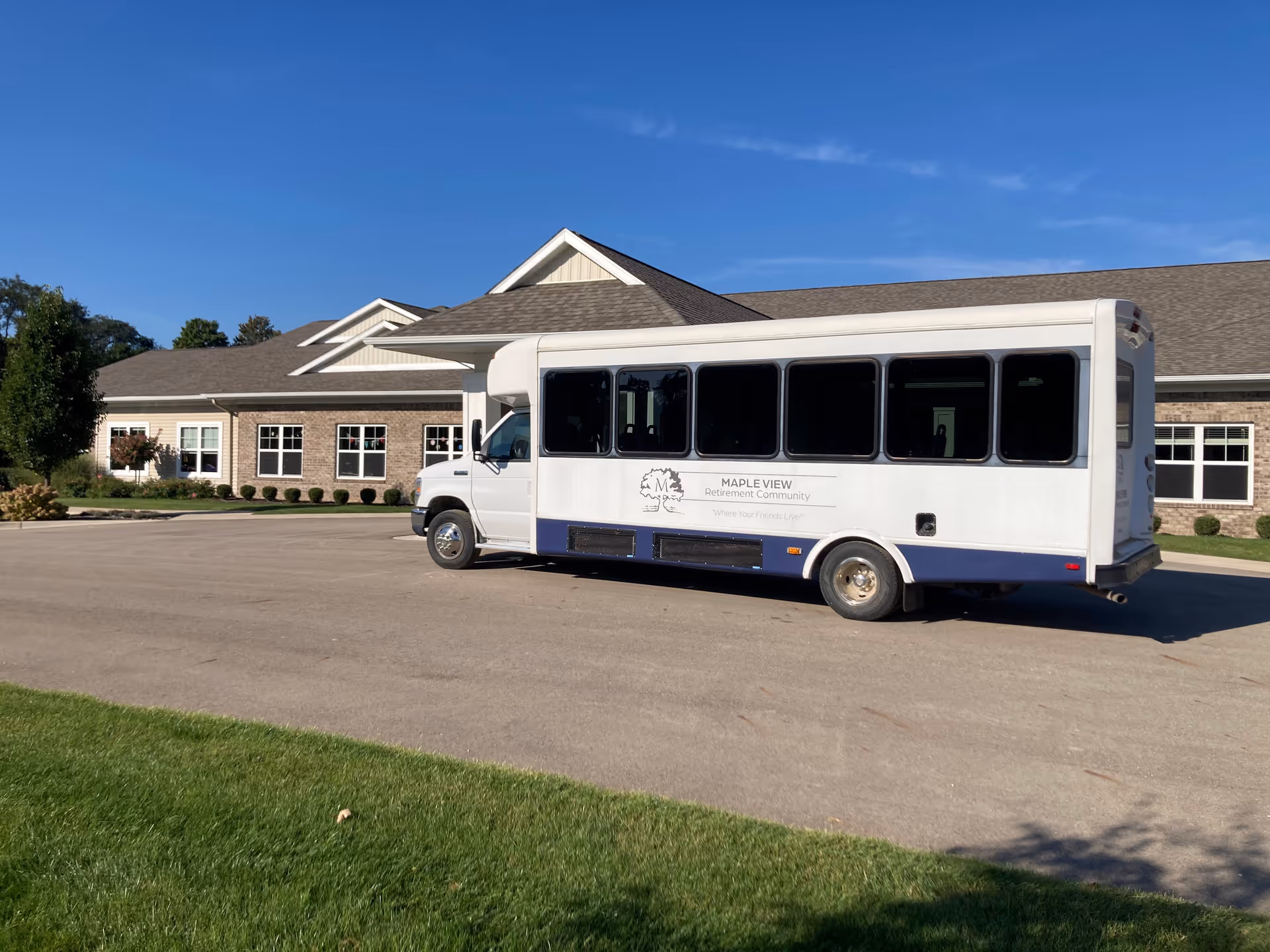 White Maple View Retirement Community shuttle parked in front of the facility's single-story building under a clear blue sky.