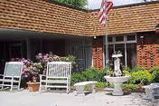 Outdoor seating area in front of a brick building with white benches, stone tables, potted plants, flowers, and an American flag on a flagpole.