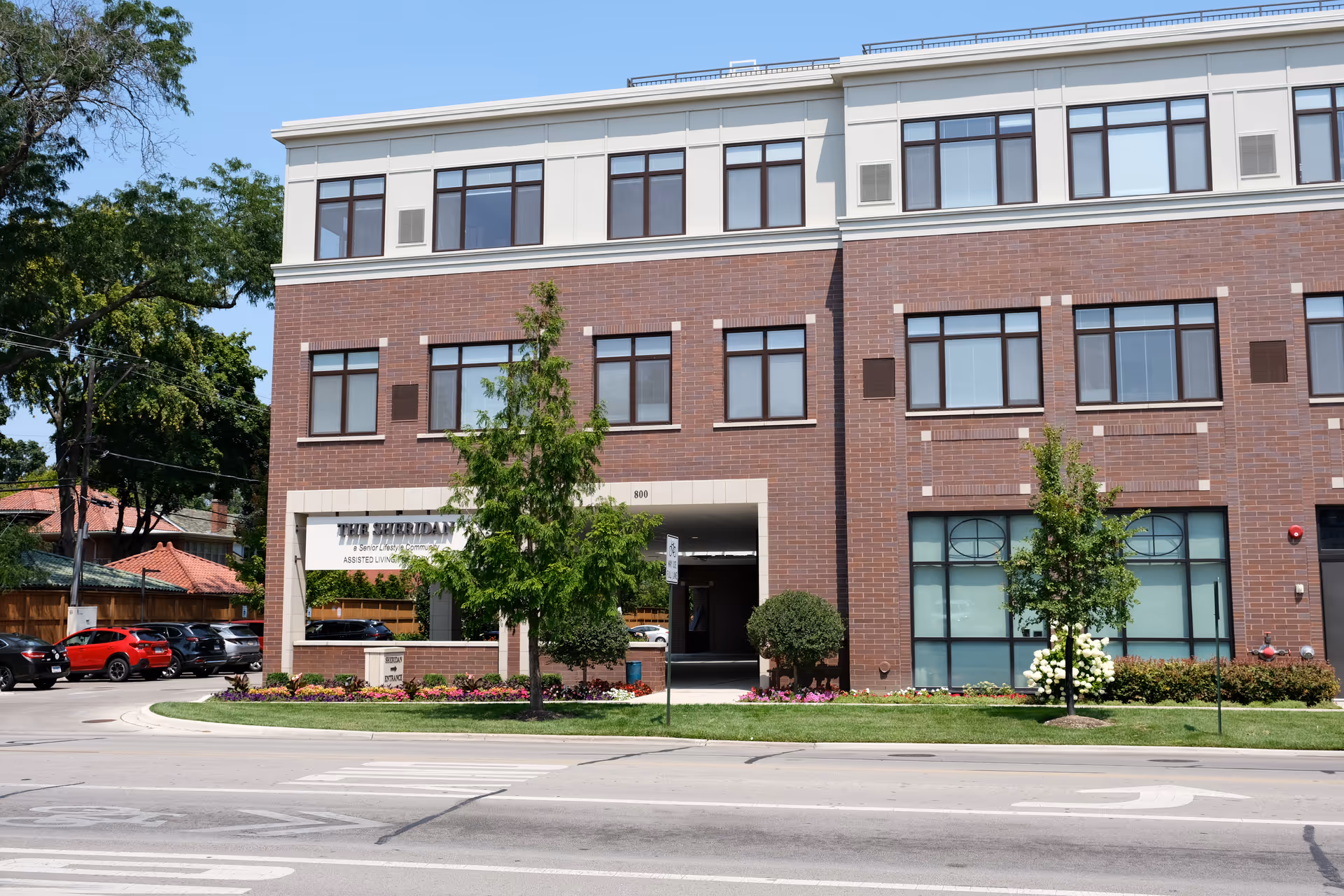 Front exterior of The Sheridan at River Forest, a three-story brick building with an entrance, landscaping, and parked cars along the street.