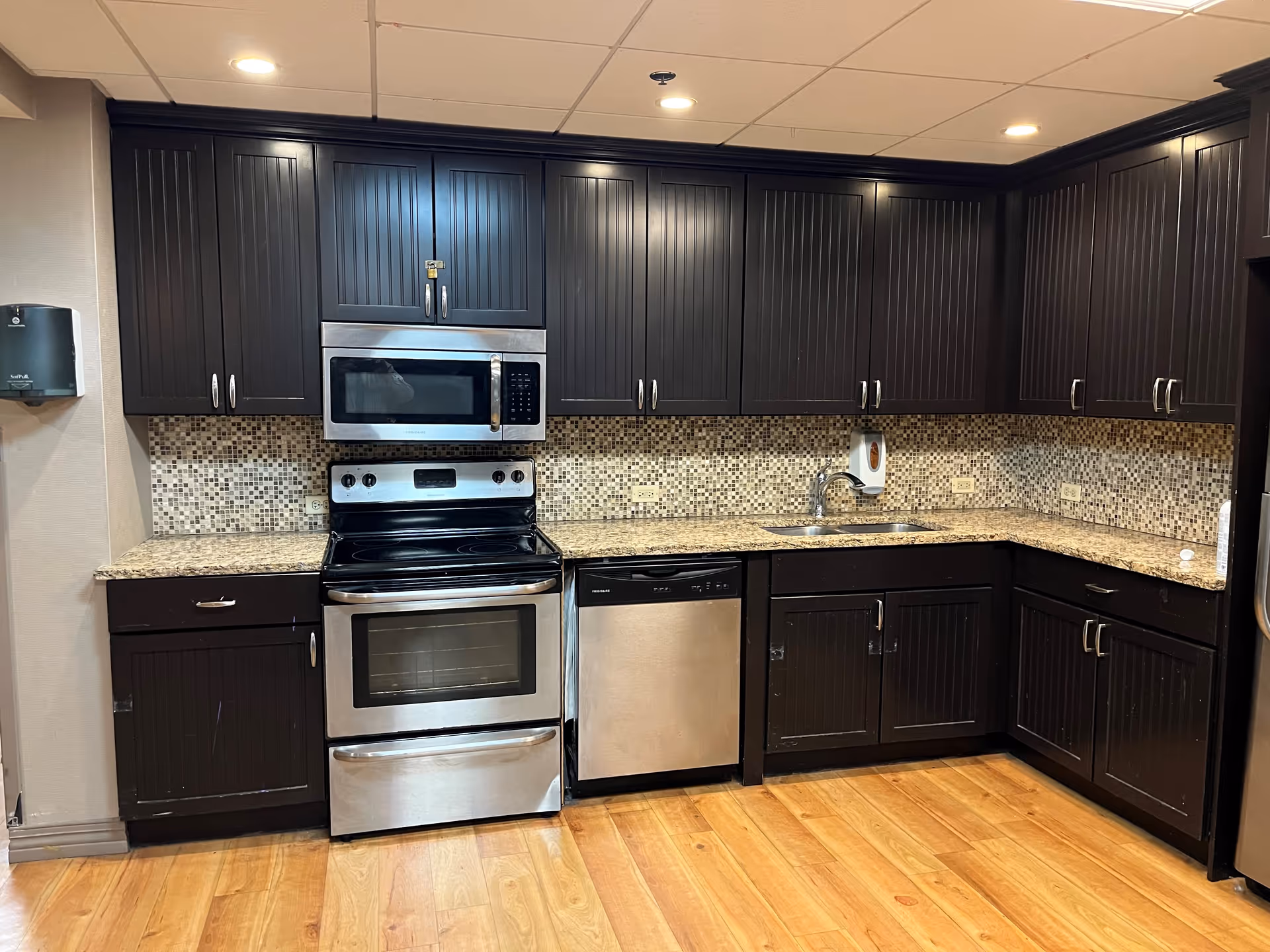 A modern kitchen with dark wood cabinets, a mosaic tile backsplash, granite countertops, a stainless steel oven with a stove, a microwave above it, a dishwasher, and a sink. The floor is wooden and the ceiling has recessed lighting.